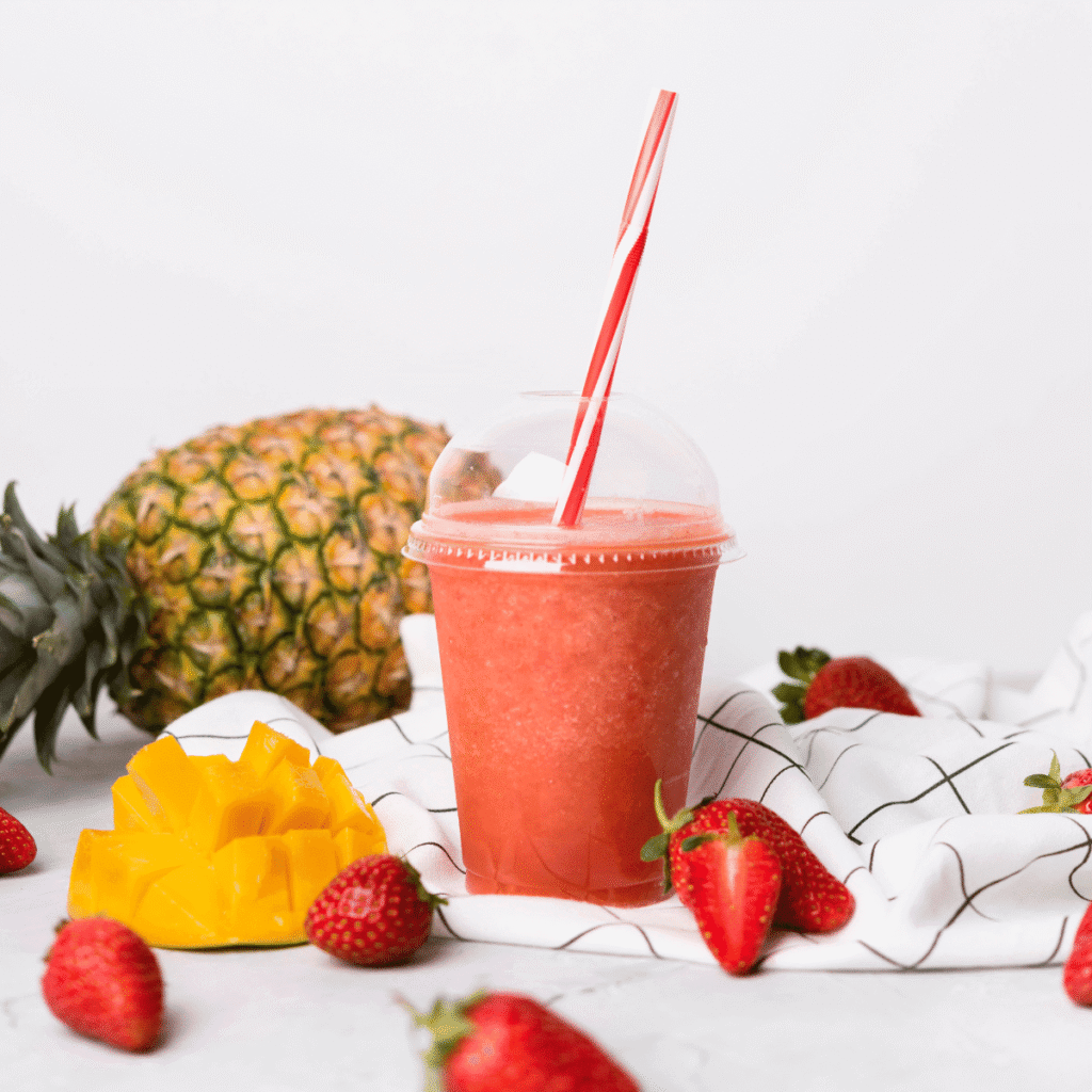 Upclose shot of smoothie filled lidded cup surrounded by pineapple, strawberries and a mango