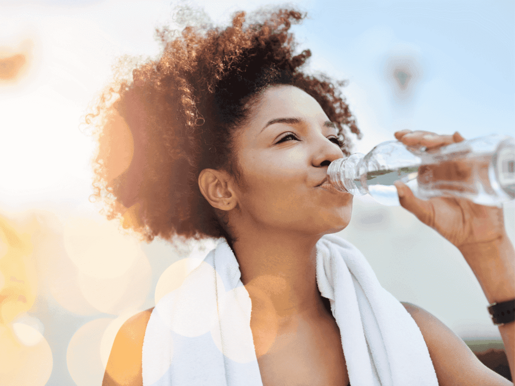 Woman with curly hair drinking water, she is outside and has a towel around her neck