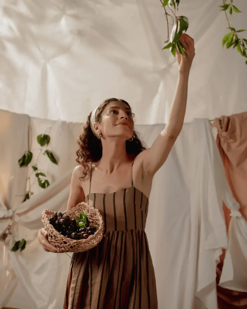 Smiling-woman-with-a-basket-of-grapes-in-one-hand-raising-her-other-hand-to-touch-leaves