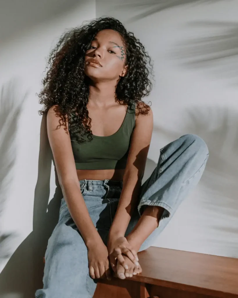 slim-black-woman-with-curly-hair-sitting-on-a-wooden-bench-indoors.-Shadows-of-plants-behind-her-on-a-plain-grey-wall.-Pillow-Method