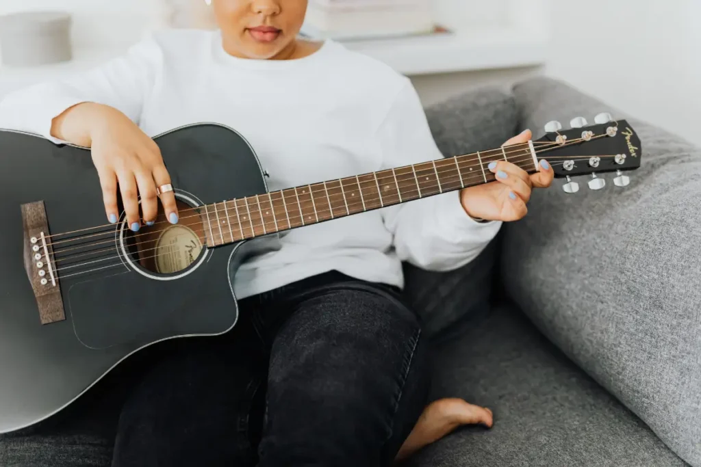 A woman in a white sweater strumming an acoustic guitar while sitting comfortably on a sofa. Hobbies For Women In Their 20s