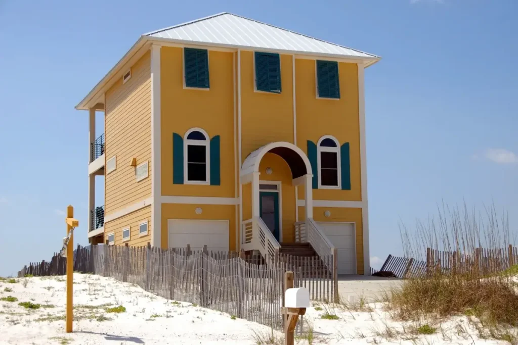 A vibrant yellow beach house with green shutters sits on a sandy dune, surrounded by a fence. Funny House Names