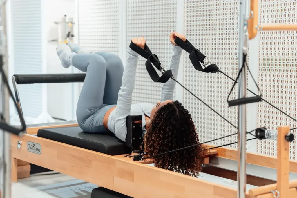 A woman wearing a gray tracksuit exercises on a Pilates reformer in a modern studio setting. Hobbies for Women Over 40