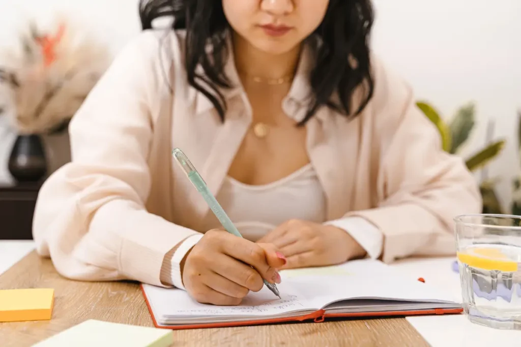 A young woman writes in her notebook while sitting at a wooden table indoors.