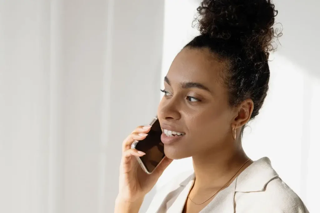 Smiling businesswoman with a hair bun talking on the phone indoors, holding a glass. Self-Care Bingo