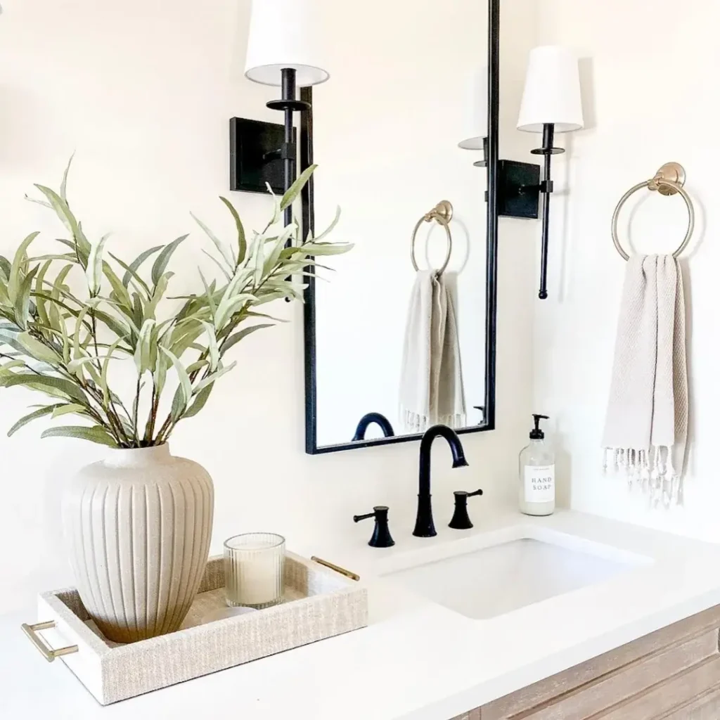 Modern white bathroom vanity with black framed mirror, black wall sconces, brass towel rings, and a linen tray with a ceramic vase of eucalyptus