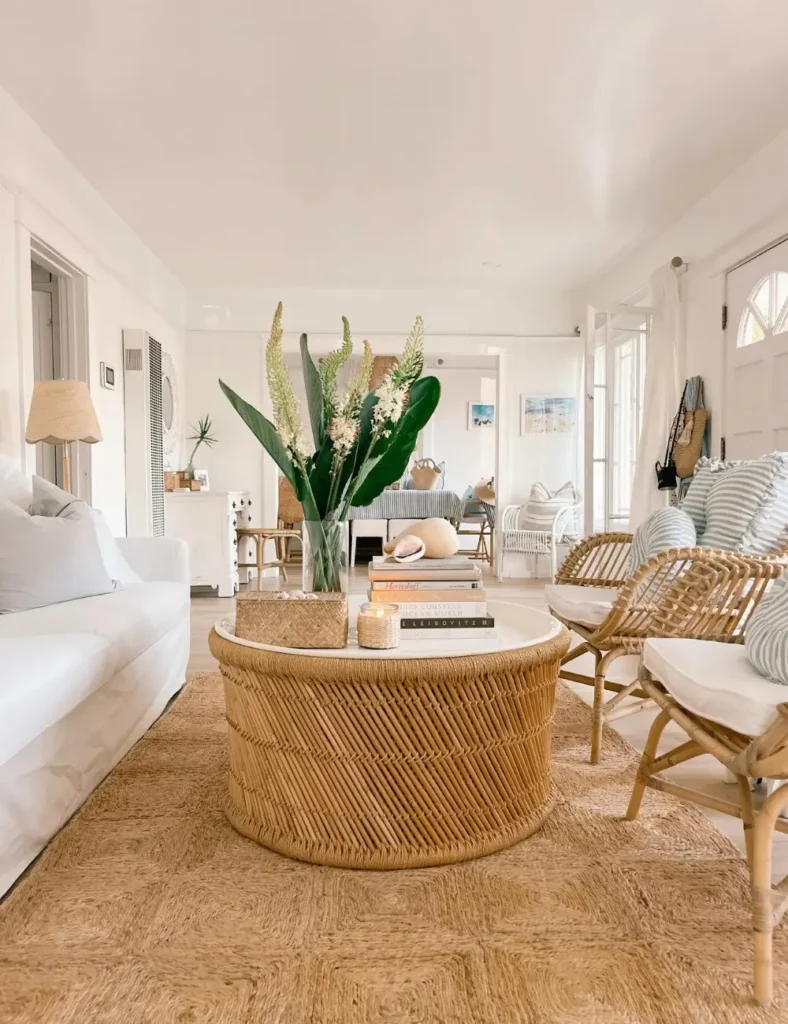 White coastal living room with a round rattan coffee table, jute rug, and rattan accent chairs styled with tropical greenery and books.