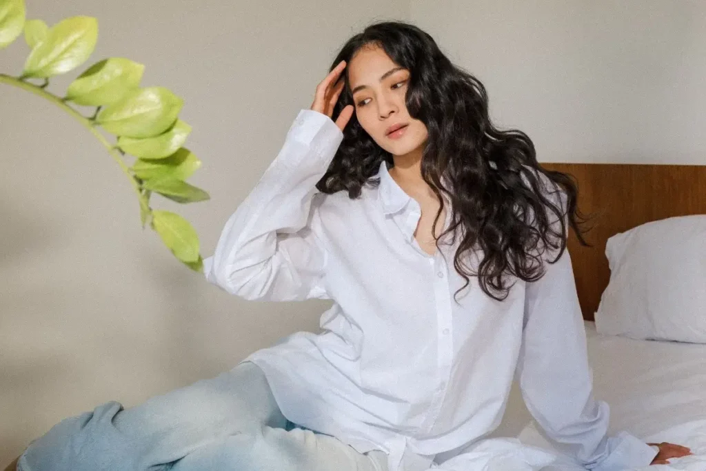 Woman in an oversized white button-down sitting on a bed, running a hand through her hair beside a trailing green plant.