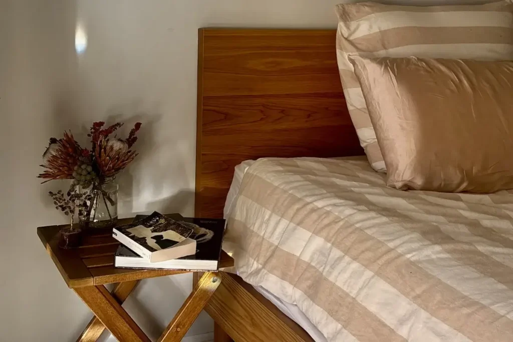 Wooden nightstand with a dried flower arrangement and a stack of books beside a bed with striped tan linens.