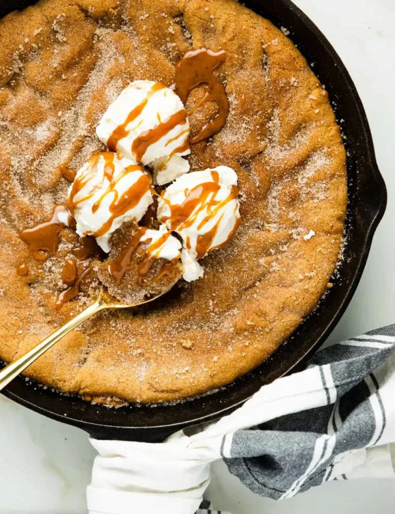 Overhead shot of a giant cinnamon sugar cookie in a cast iron skillet, topped with scoops of vanilla ice cream and a caramel drizzle.