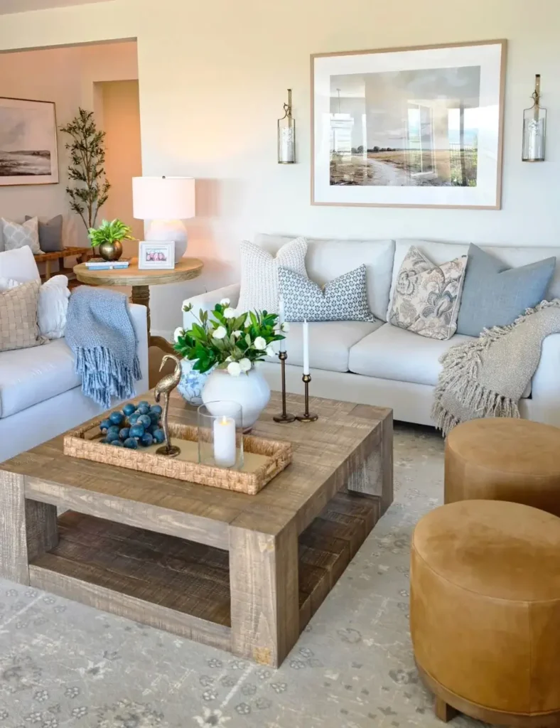 Neutral coastal living room with a wood coffee table, blue and gray accent pillows, brass candlesticks, and a framed landscape print.
