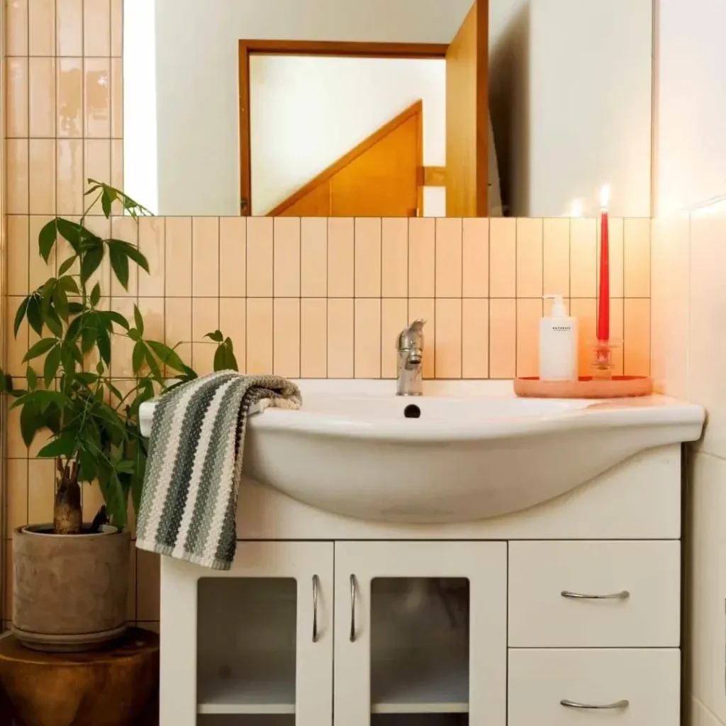 Small bathroom with peach vertical tile, white vanity, striped hand towel, potted plant, and a red taper candle on a pink tray