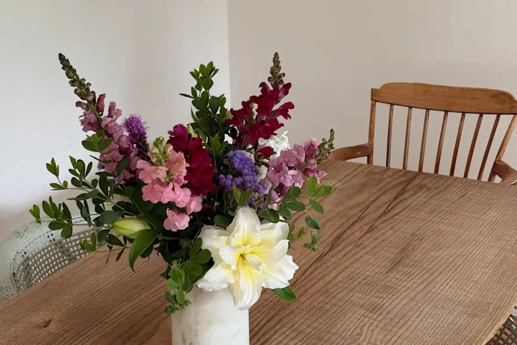 Wild-style flower arrangement of snapdragons, statice, and a white lily in a marble vase on a wooden dining table.