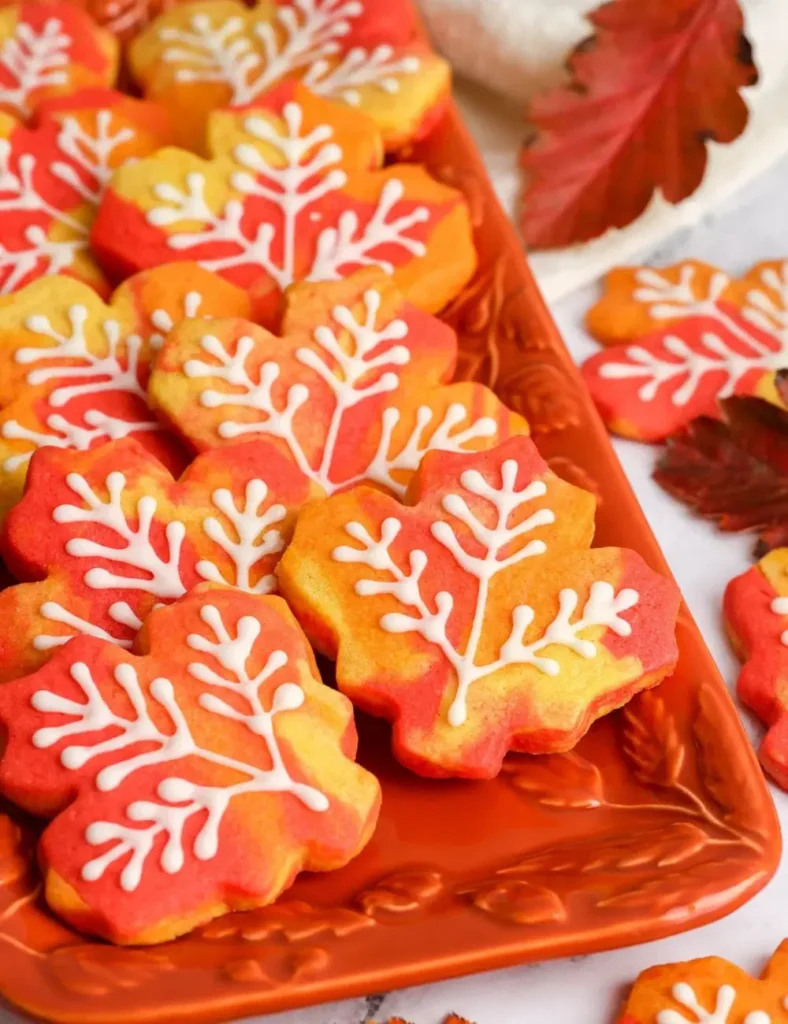 Maple leaf-shaped sugar cookies in red, orange, and yellow with white royal icing vein details, arranged on an orange platter.