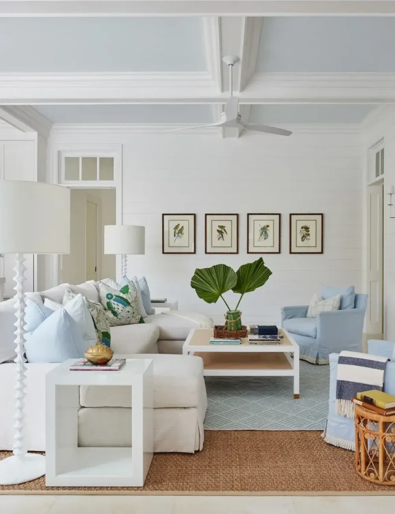 Bright coastal living room with a pale blue ceiling, white beamed trim, blue slipcovered armchair, and a seagrass rug.