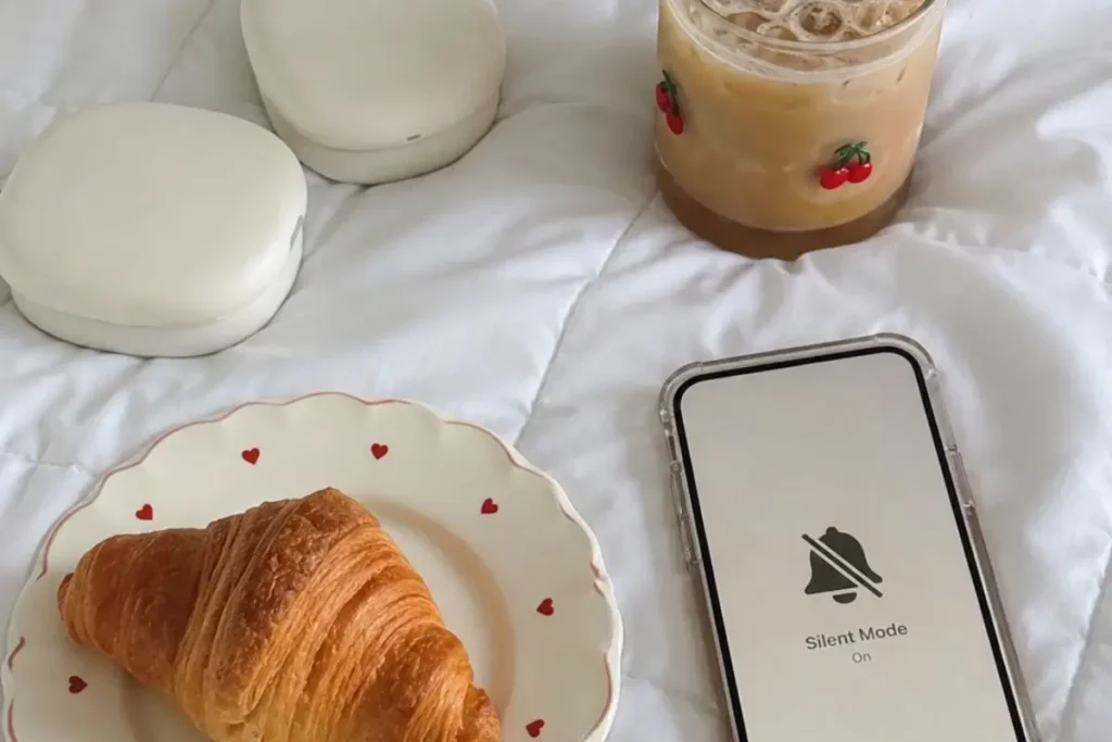 Silent Mode phone next to a flaky croissant on a heart-patterned plate and an iced coffee in a cherry-print glass, styled on white bedding.