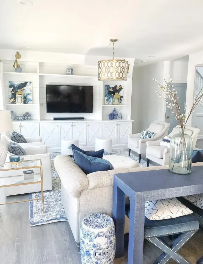 Coastal living room with built-in white shiplap shelving, blue and white ginger jars, a chinoiserie garden stool, and a brass pendant light.