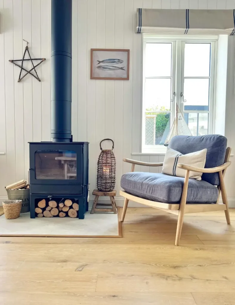 Cottage-style coastal living room with a navy wood-burning stove, wood-paneled walls, a woven lantern, and an oak armchair with a blue cushion.