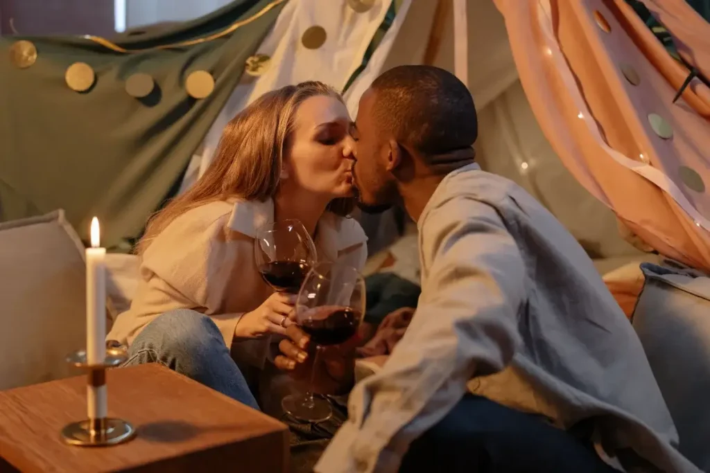 A couple shares a romantic kiss while holding wine glasses in cozy indoor setting. Date Night Ideas