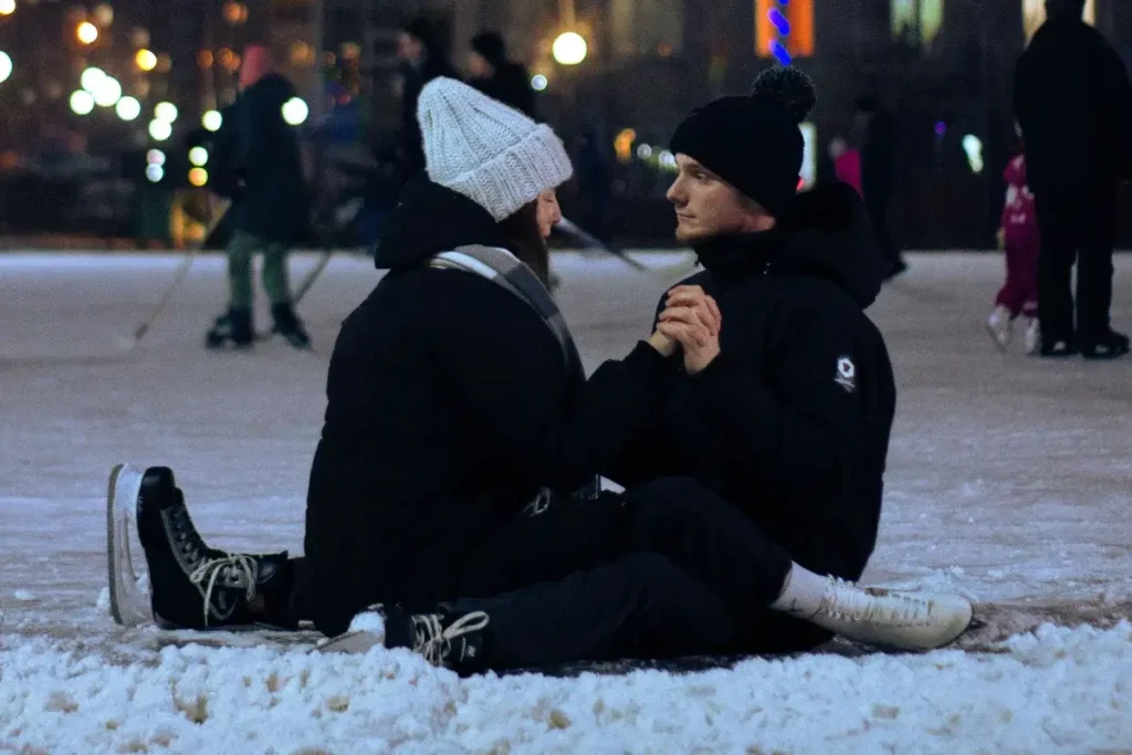 A couple sitting together on snowy ground at a lively outdoor ice rink at night. Date Night Ideas
