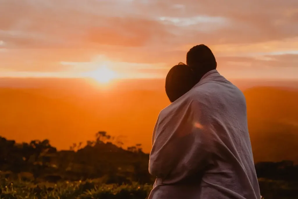 A couple wrapped in a blanket watches the warm sunset from a hilltop, conveying intimacy and serenity. Date Night Ideas