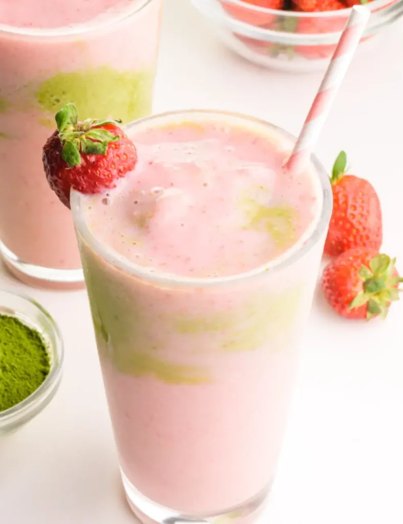 A glass of layered pink and green strawberry matcha smoothie beside fresh strawberries and a bowl of matcha powder