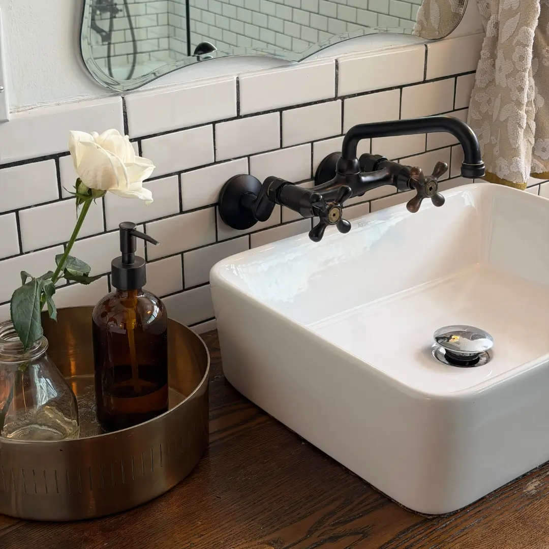 White vessel sink on a dark wood counter with a black wall-mounted faucet, white subway tile, and an amber glass soap dispenser on a brass tray