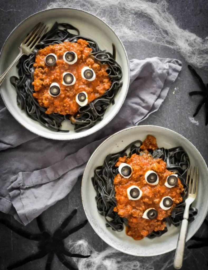 Black squid ink pasta topped with beef ragu and mozzarella and olive eyeballs in a white bowl with plastic spiders