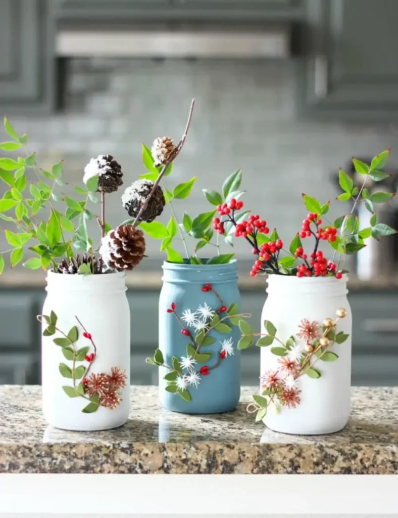 Chalk-painted mason jar vases decorated on the outside with faux winter greenery and berries, displayed together on a table.