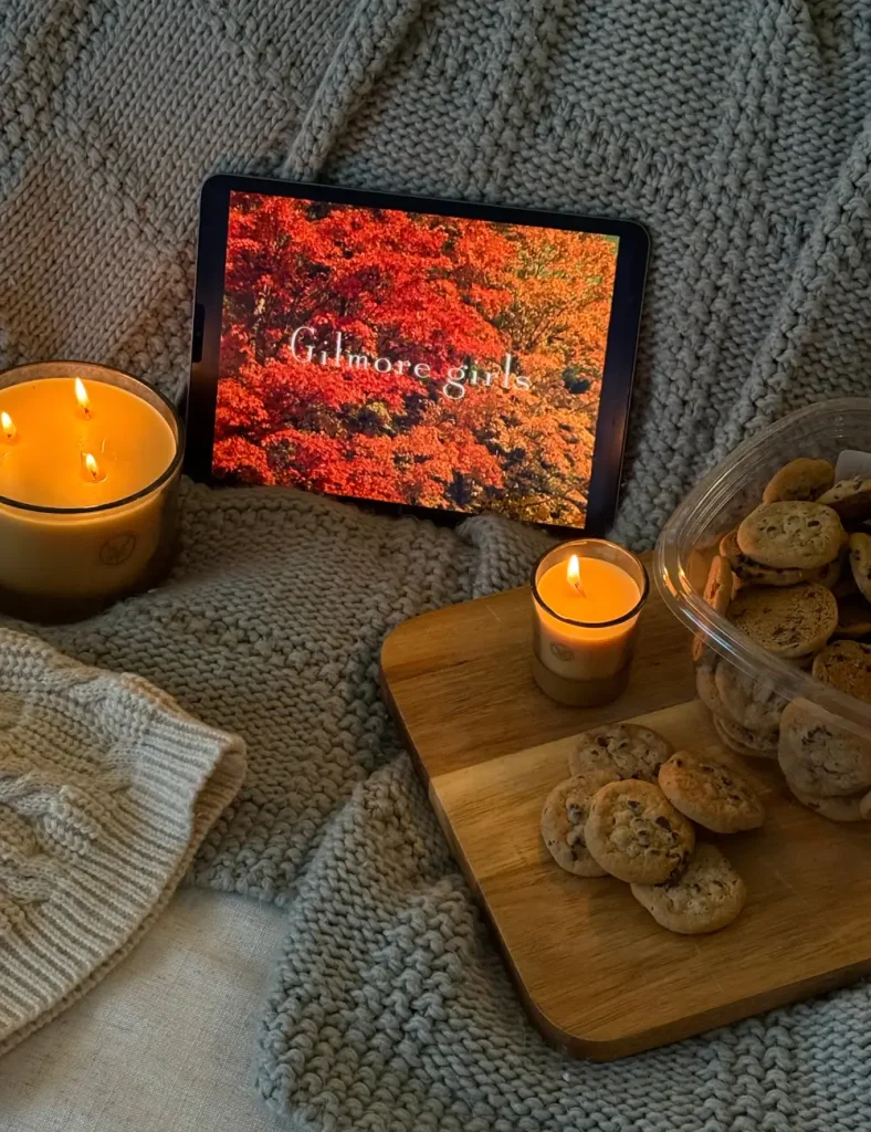 Cozy scene with lit candles, chocolate chip cookies on a wooden board and a tablet on a chunky knit blanket