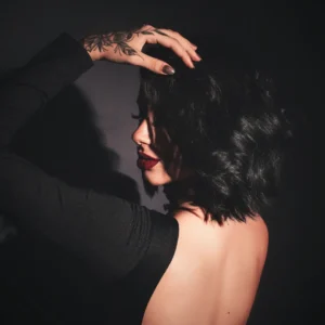 Woman with a dark bob and red lipstick in low light, turned away from the camera with her hand in her hair