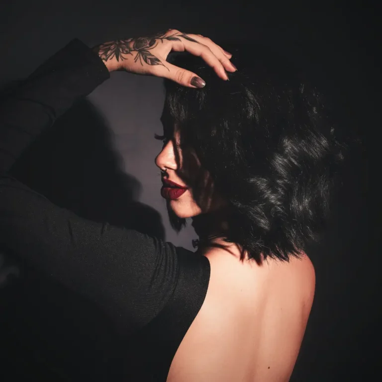 Woman with a dark bob and red lipstick in low light, turned away from the camera with her hand in her hair