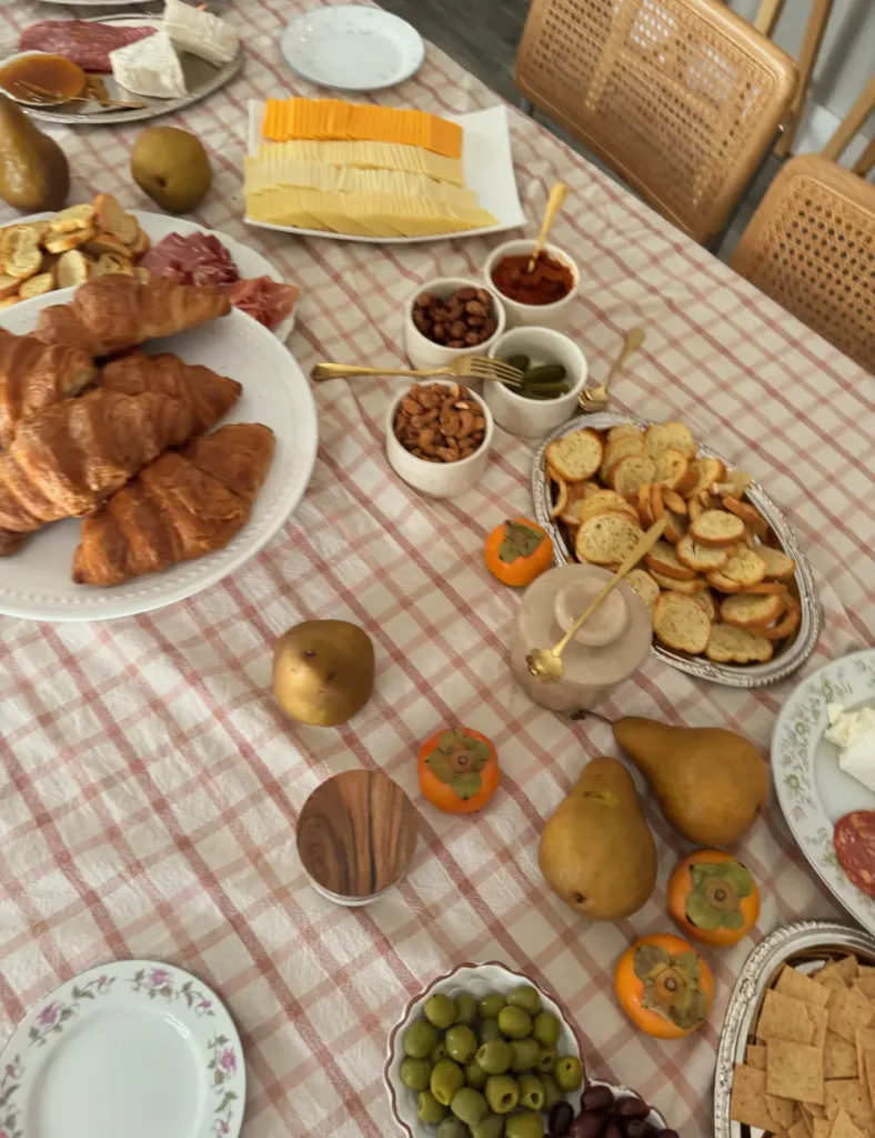 Fall grazing table on a pink gingham cloth with croissants, sliced cheeses, persimmons, pears, olives and small bowls of nuts