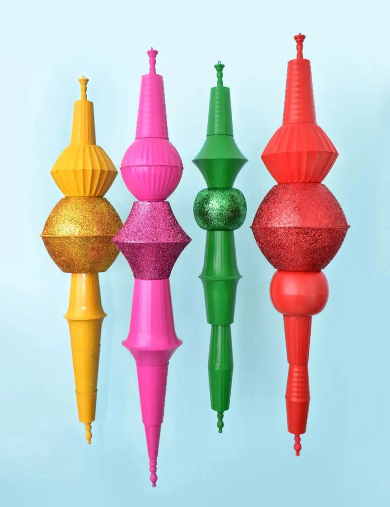Four oversized finial Christmas ornaments in pink, red, green and gold leaning against a wall.