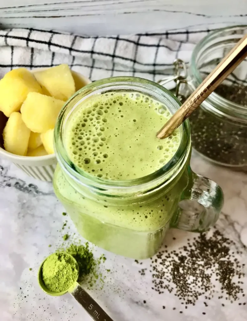 Green pineapple matcha smoothie beside matcha powder, fresh pineapple chunks, and chia seeds