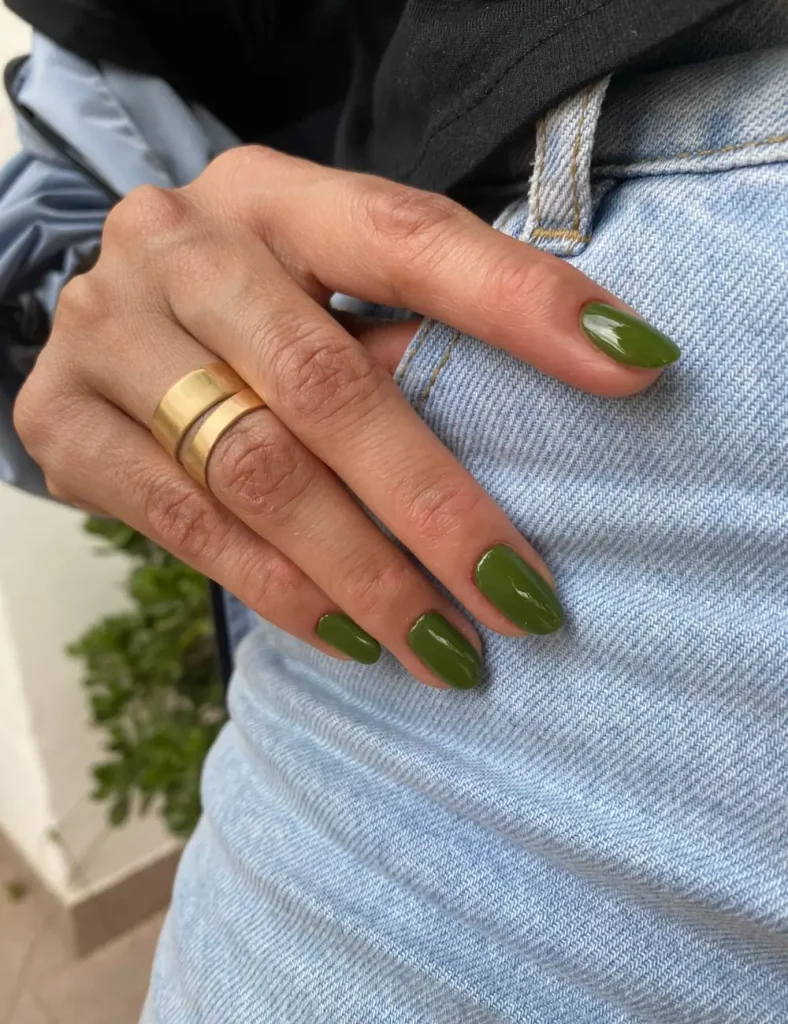 Hand with olive green almond-shaped nails and two stacked gold rings resting on light blue denim