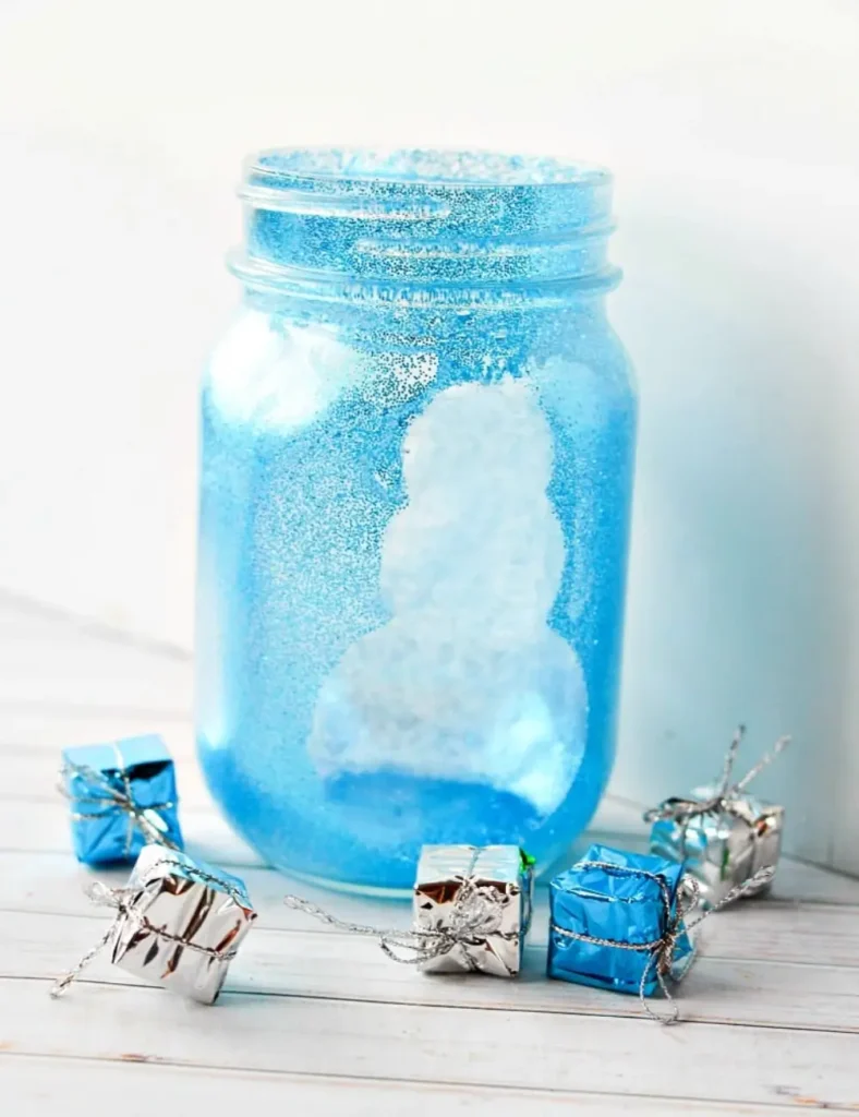Mason jar coated in sparkly glitter with a clear snowman silhouette in the center, glowing from a candle inside.