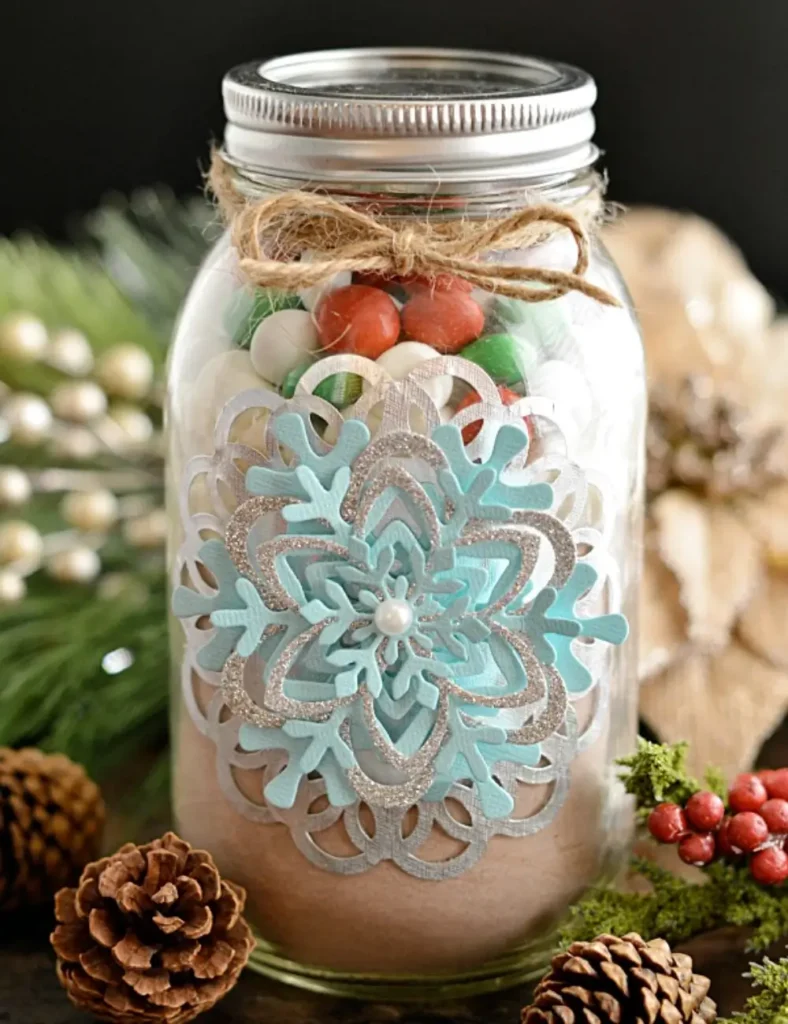 Mason jar decorated with a silver doily and paper snowflakes, tied with twine and filled with hot cocoa mix, marshmallows and mint candies.