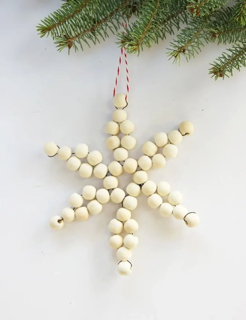 Natural wood bead snowflake ornament strung on red and white striped twine on a christmas tree, hanging against a pale gray background.