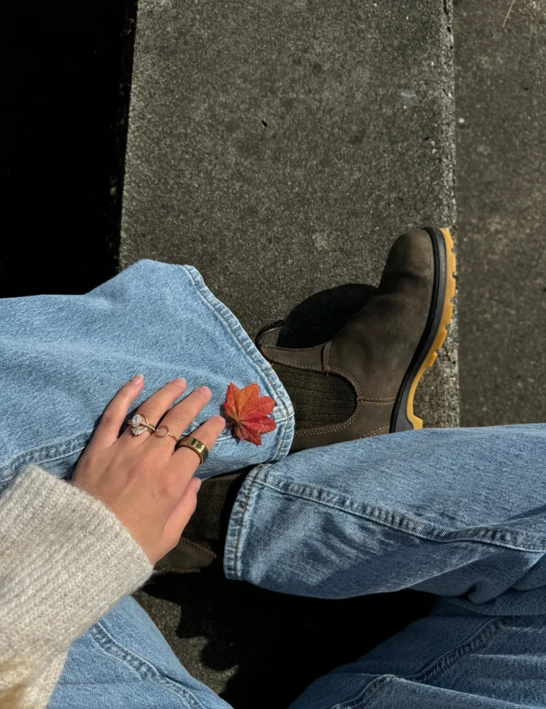 Overhead view of a person sitting on a step in light jeans and brown Chelsea boots with a red maple leaf resting on their knee