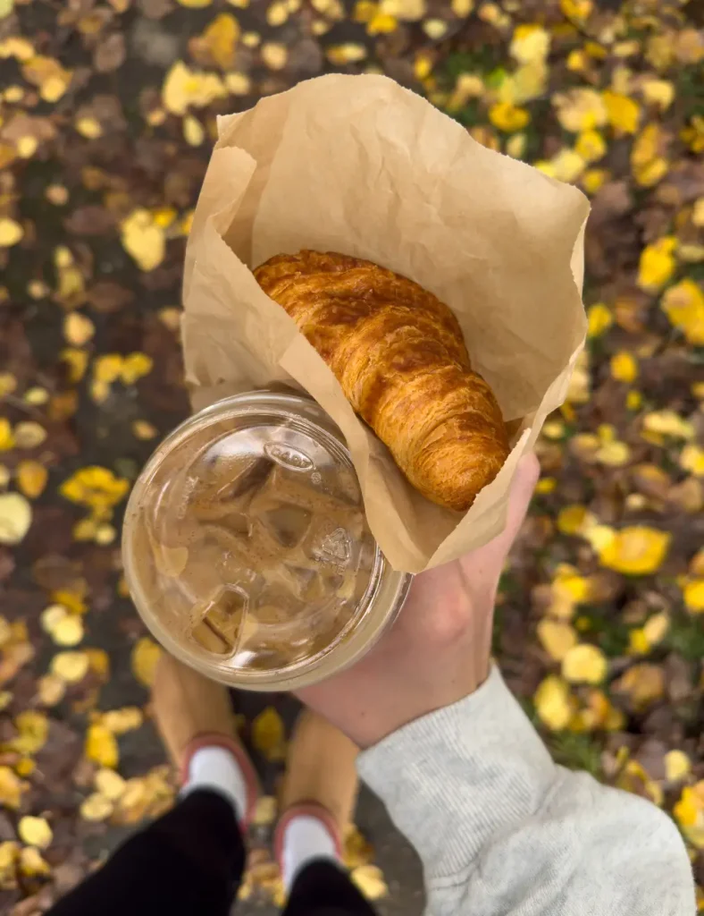 Overhead view of hands holding an iced coffee and a croissant in paper above a path scattered with yellow leaves