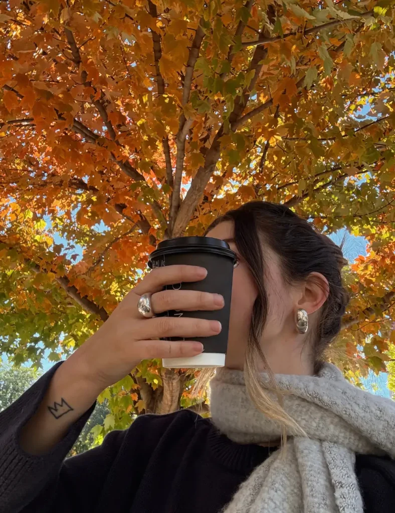 Person in a black sweater and cream scarf drinking from a black takeaway cup under a bright orange maple tree