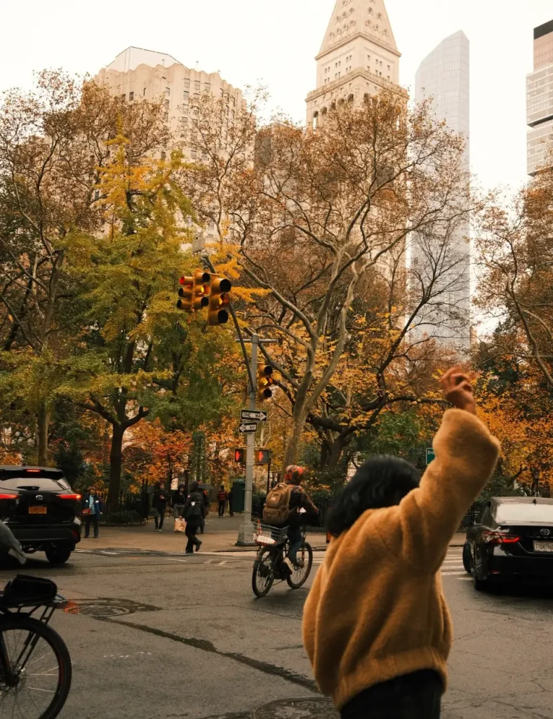 Person in a camel teddy coat hailing a cab at a city crosswalk with yellow and orange autumn trees and tall buildings behind
