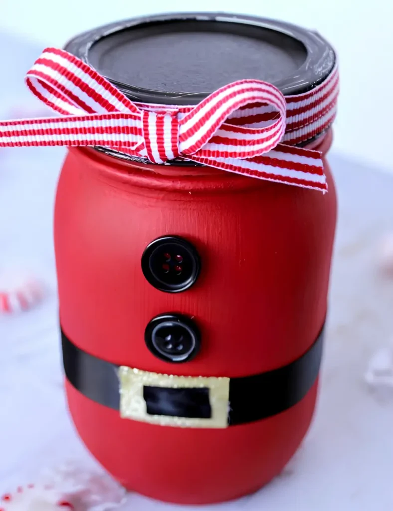 Red painted mason jar decorated like Santa with a black ribbon belt, gold buckle and two black buttons, with a striped ribbon bow on the lid.