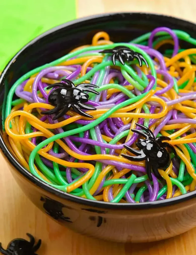 Spaghetti dyed orange, purple, and green twisted together in a white bowl with a plastic spider on the rim