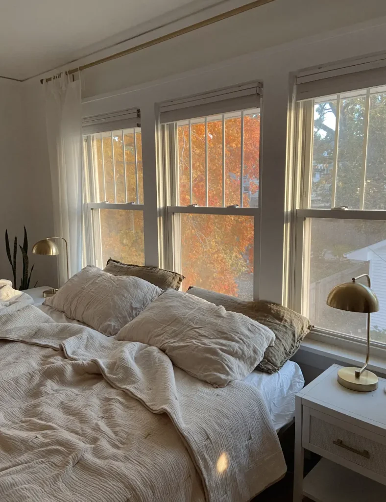 Sunlit bedroom with rumpled linen bedding in oatmeal and taupe, brass table lamps and three windows showing orange fall foliage