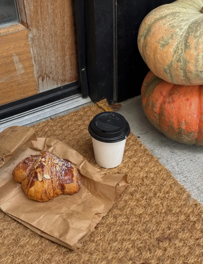 Takeaway coffee cup and almond croissant on a paper bag on a coir doormat beside two large heirloom pumpkins