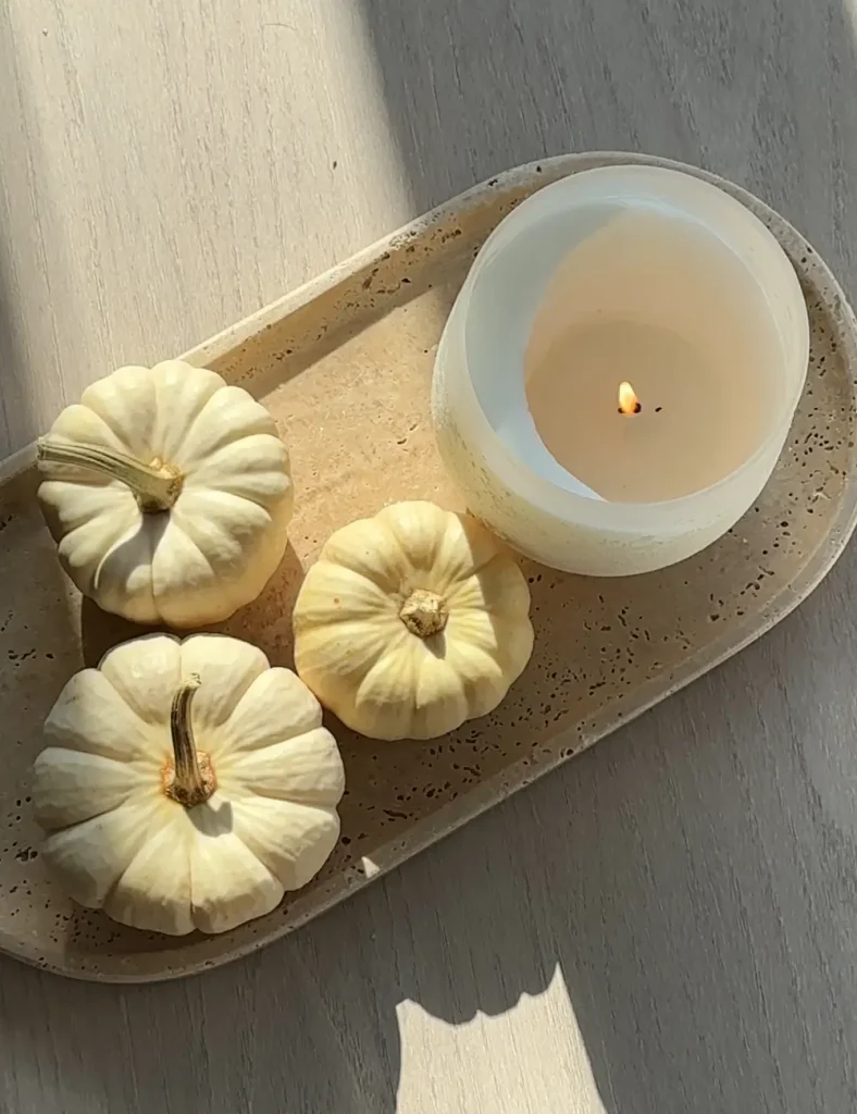 Three small white pumpkins and a lit candle arranged on an oval travertine tray in afternoon sunlight