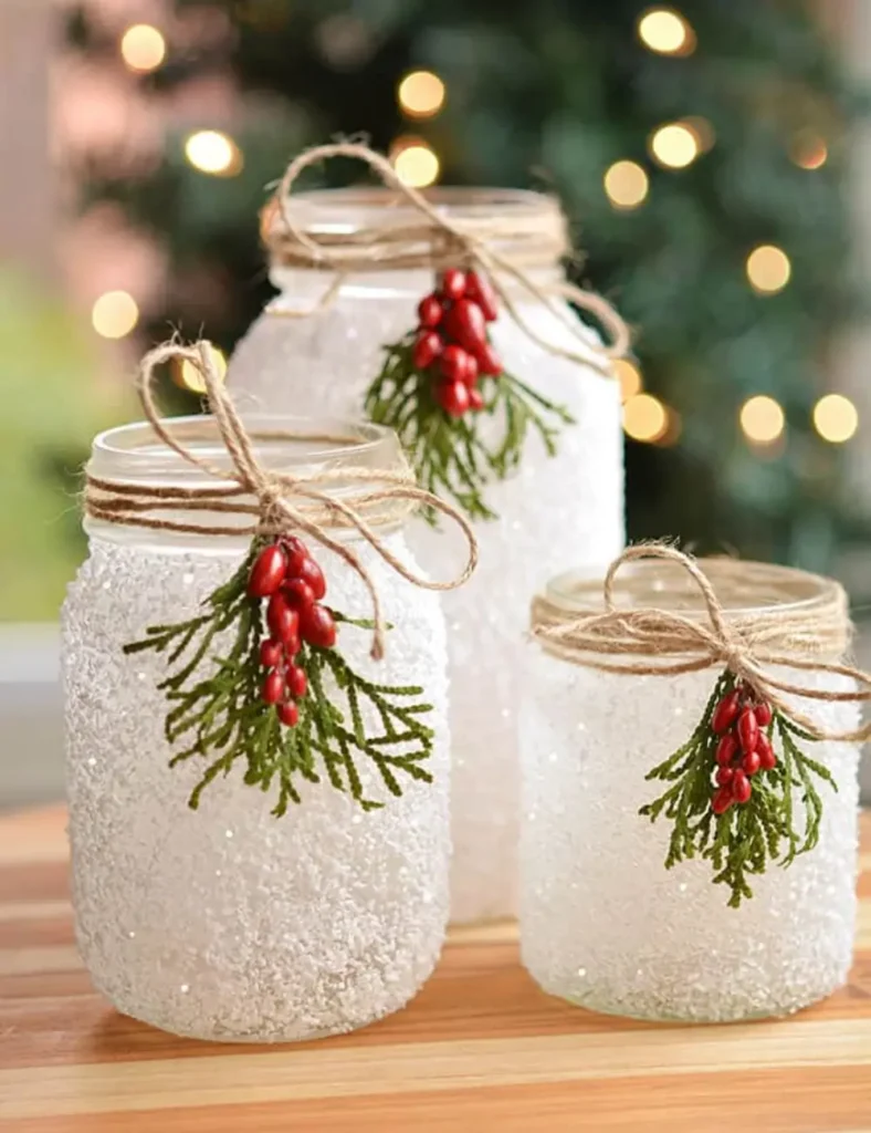 Three snowy mason jar luminaries covered in Epsom salt “snow,” decorated with greenery, berries and twine, glowing with battery tea lights.