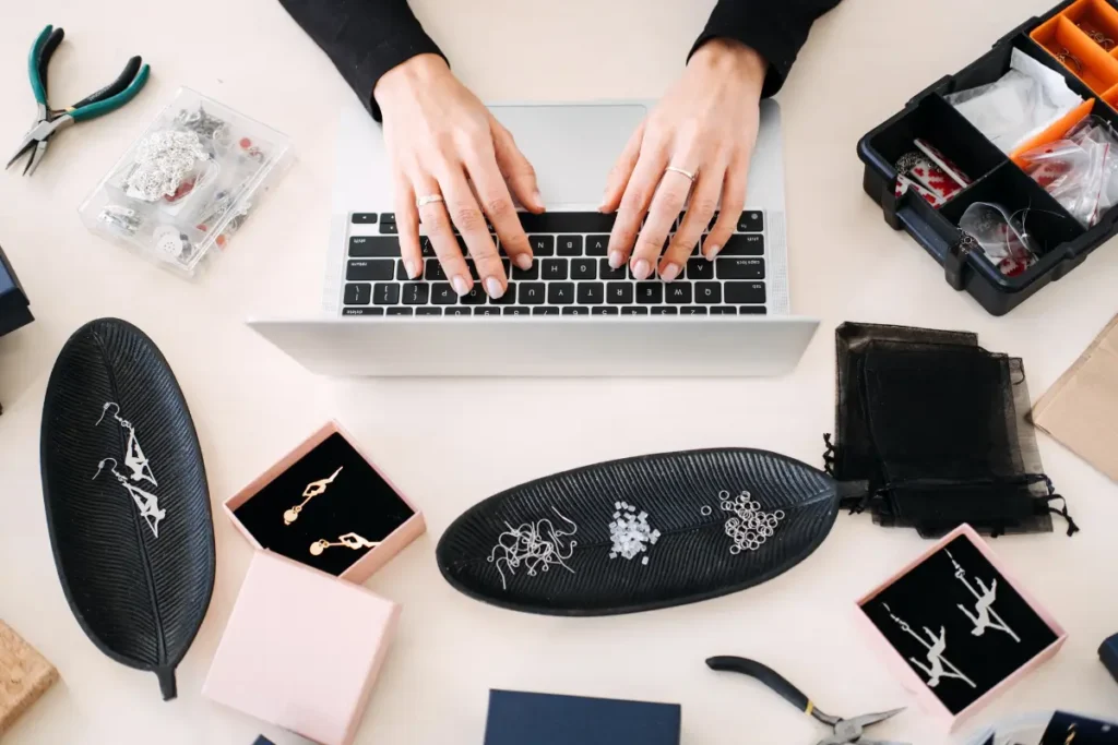 Jewelry maker setting up her online shop on a laptop, surrounded by her handmade pieces.
